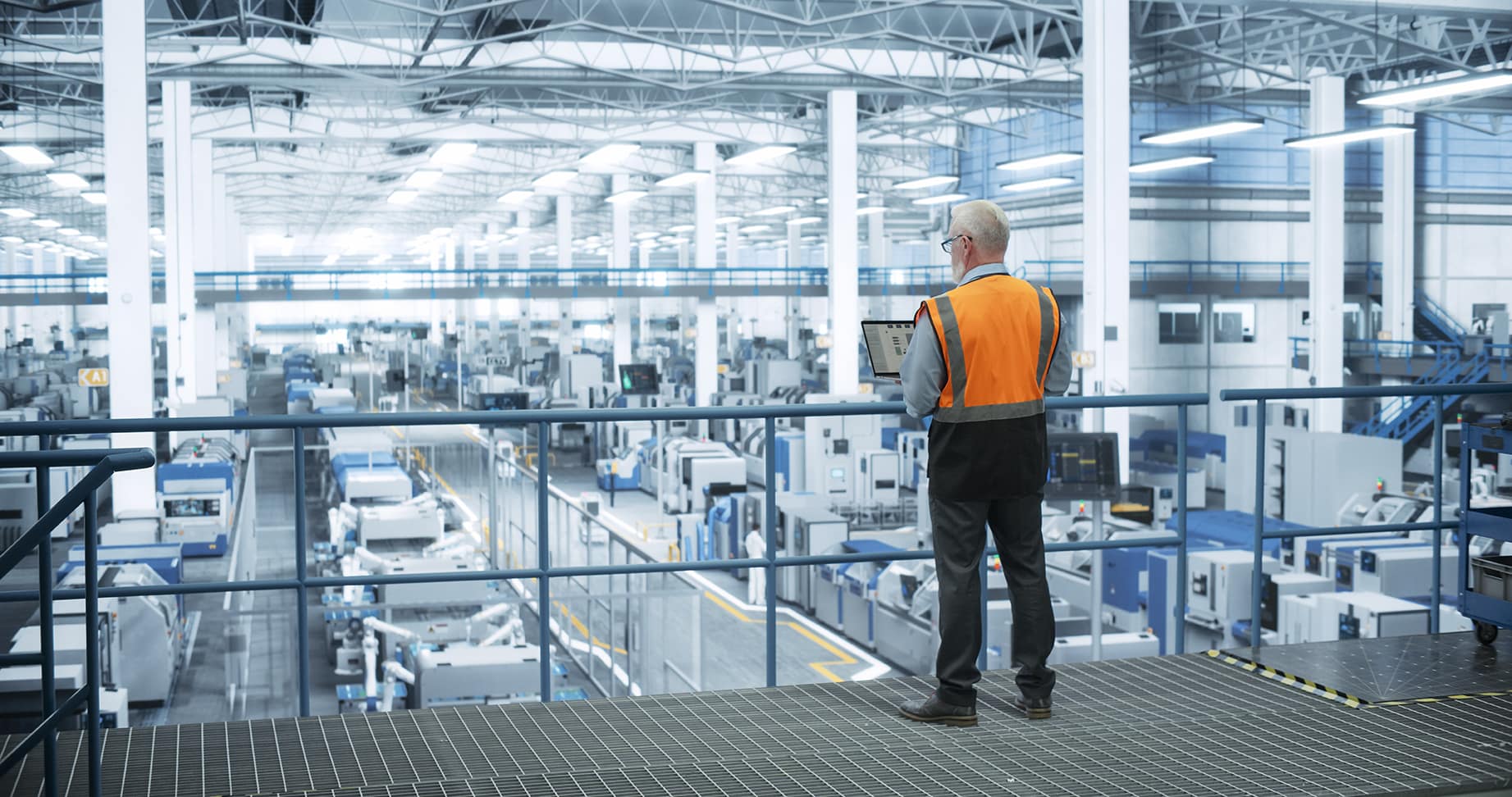 Industrial Engineer Working at a Technology Factory. Middle Aged Male Using Laptop Computer at Work, Browsing Electronics Information Online. Man’s Back in Safety Vest Turned to Camera